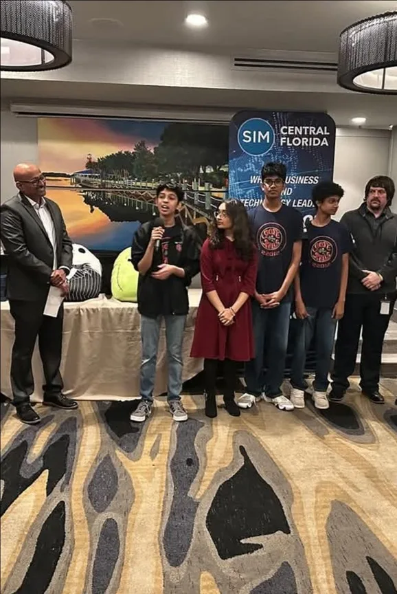 Monish speaking into a microphone with robotics teammates at a SIM Central Florida event