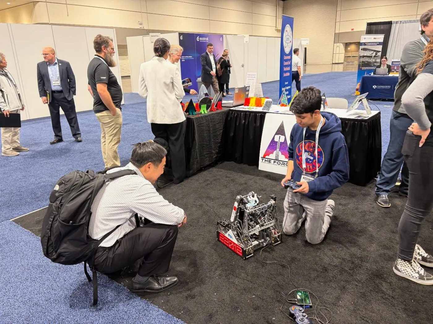 Monish kneeling beside a competition robot on the SPACECOM expo floor as visitors watch